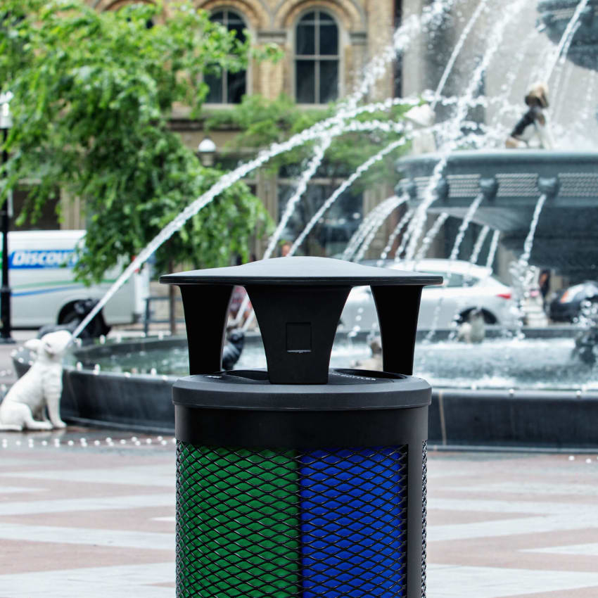 black metal outdoor waste and recyclables container with a plastic canopy lid at a city park with a water fountain