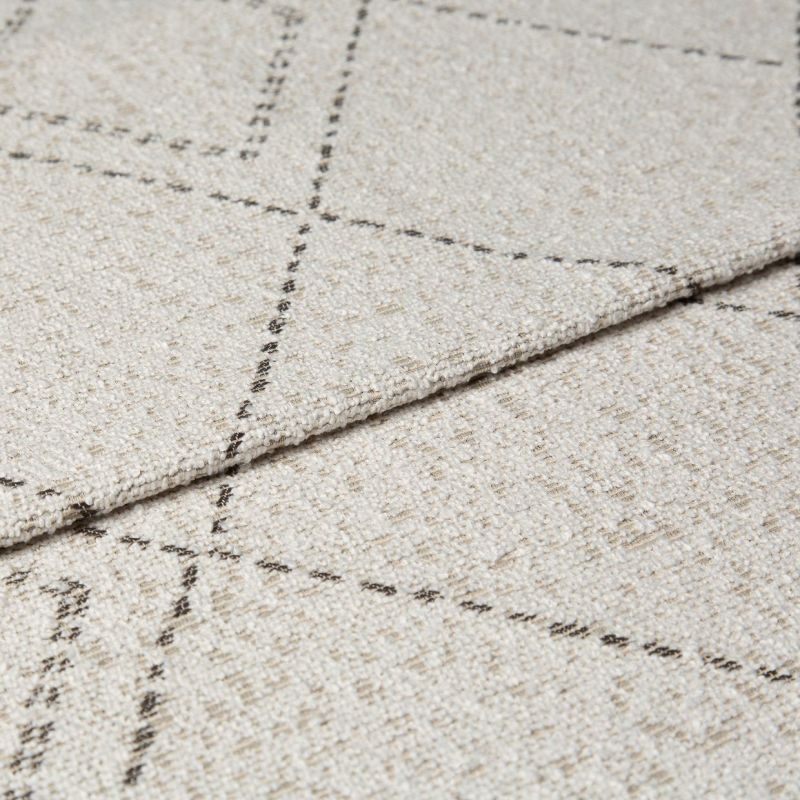Beige woven rug with thin dashed dark grid lines, folded at the edge to show layered thickness and looped texture; close-up studio shot with neutral lighting on a flat surface.