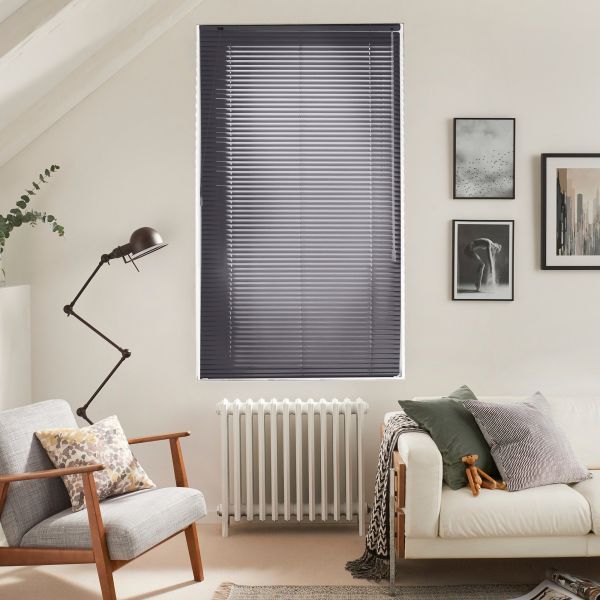 Gray horizontal Venetian blinds covering a tall central window, closed and filtering soft daylight; slender radiator beneath; neutrally furnished living room with armchair, sofa, framed art, and lamp.