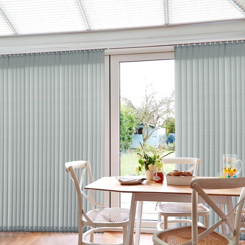 Pale-gray vertical fabric blinds with narrow slats covering a sliding glass door, partially open to reveal a sunlit garden; dining table and chairs with vase and pastries in the foreground.