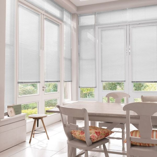 White horizontal Venetian blinds lowered across tall bay windows, slats mostly closed and softly filtering daylight, in a bright dining sunroom with white table, chairs, cushions and garden view.