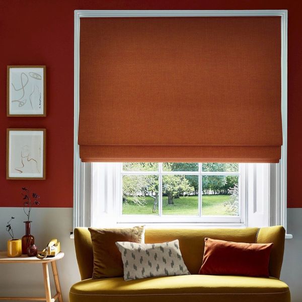 Burnt-orange Roman blind, lowered to the sill with soft folds; blocking light and framing a white-paned window; living room with mustard sofa, cushions, red walls, framed sketches and garden view.