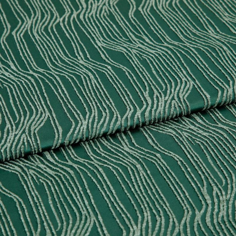 Green jacquard fabric with raised cream wavy lines, folded and overlapping; pattern runs diagonally across a close-up, evenly lit textile surface.