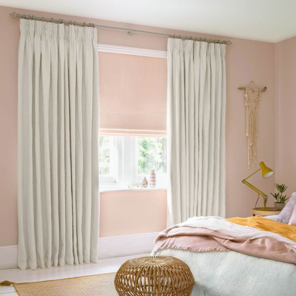 Floor-length cream pleated curtains on a decorative rod, framing a lowered blush roller blind that softly filters daylight into a pastel-pink bedroom with bed, rattan stool, and angled brass lamp.