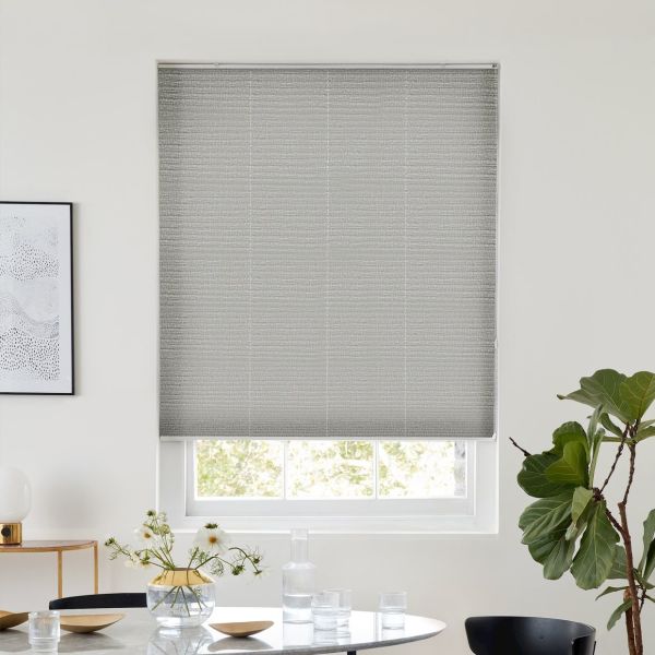 Grey textured roller blind fully lowered, blocking most light; minimalist dining nook with round table, glassware and vase, potted fiddle‑leaf fig, soft daylight filtering around blind edges.