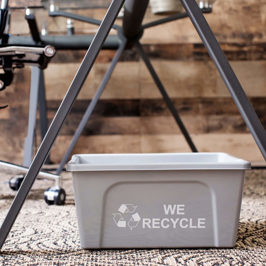 grey plastic deskside paper recycling container with we recycled printed on the side sitting on the floor in an office