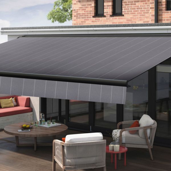 A large retractable fabric awning (dark gray, subtle diagonal stripes, front valance) mounted above sliding glass doors, extended and shading the patio; modern brick-backed outdoor seating area in daylight.