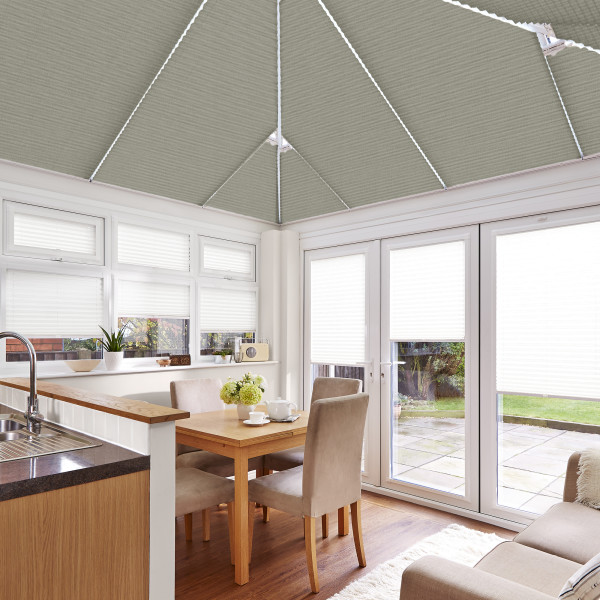 Grey pleated roof blinds with white cords, fitted across the conservatory ceiling; closed, filtering daylight over a bright kitchen-dining area with white-framed doors and windows opening to a patio garden.