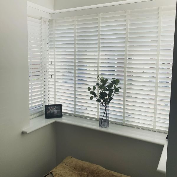 White horizontal slatted blinds covering a bay window, mostly closed and filtering soft daylight; on the windowsill a glass vase with green stems, framed photo, and sheepskin seat below.