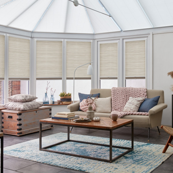 Beige pleated cellular blinds across tall conservatory windows, lowered and filtering soft daylight; a cozy sitting area with beige sofa, chunky pink throw, wooden coffee table and blue-pattern rug.