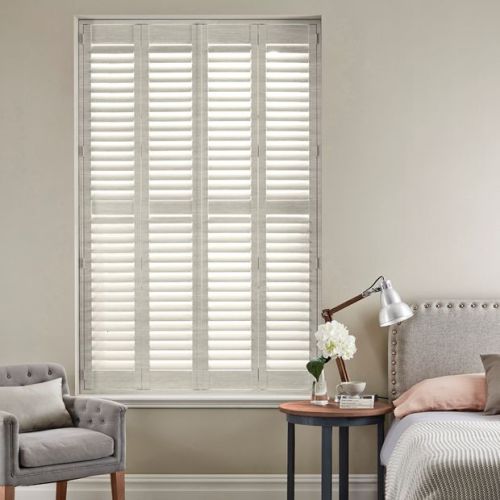 White plantation shuttersâ€”four tall louvered panelsâ€”fully closed, softly filtering daylight into a neutral bedroom corner with upholstered headboard, bedside table, lamp, vase of flowers and a gray armchair.