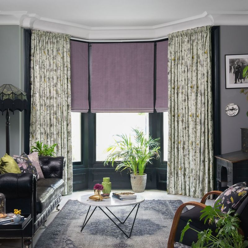 Full-length floral pleated curtains flank three mauve Roman blinds covering a bay window; blinds partially lowered, filtering daylight into a living room with dark leather sofa, coffee table, and plants.