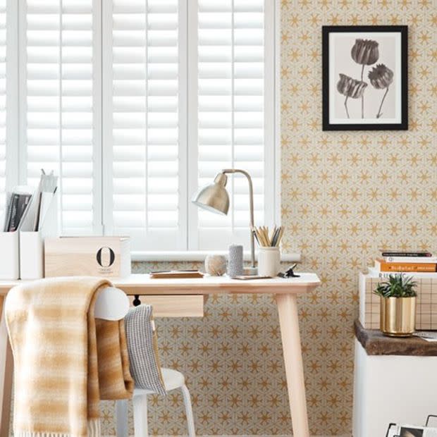 White plantation shutters over a tall window, closed with tilted slats filtering soft daylight; small home office with a light wooden desk, lamp, chair with yellow plaid throw, patterned wallpaper.