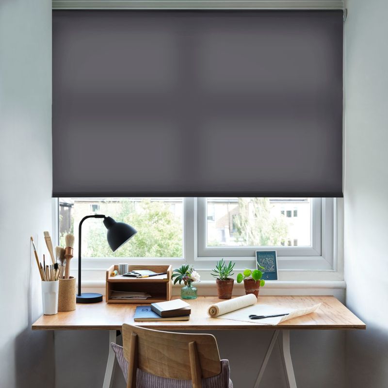 Dark grey roller blind inside the window frame, lowered almost fully and dimming daylight; above a neat wooden desk with lamp, brushes, notebooks, potted succulents, chair and treed urban view.