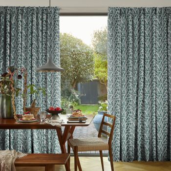 Teal-and-white patterned pleated curtains, hung on either side and slightly drawn to frame a central door, filtering daylight into a dining room with wooden table, chairs and garden view.