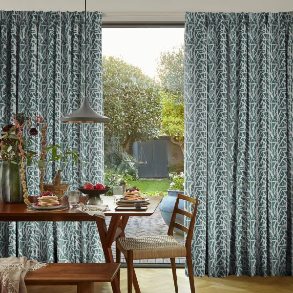 Teal-and-white patterned pleated curtains, hung on either side and slightly drawn to frame a central door, filtering daylight into a dining room with wooden table, chairs and garden view.