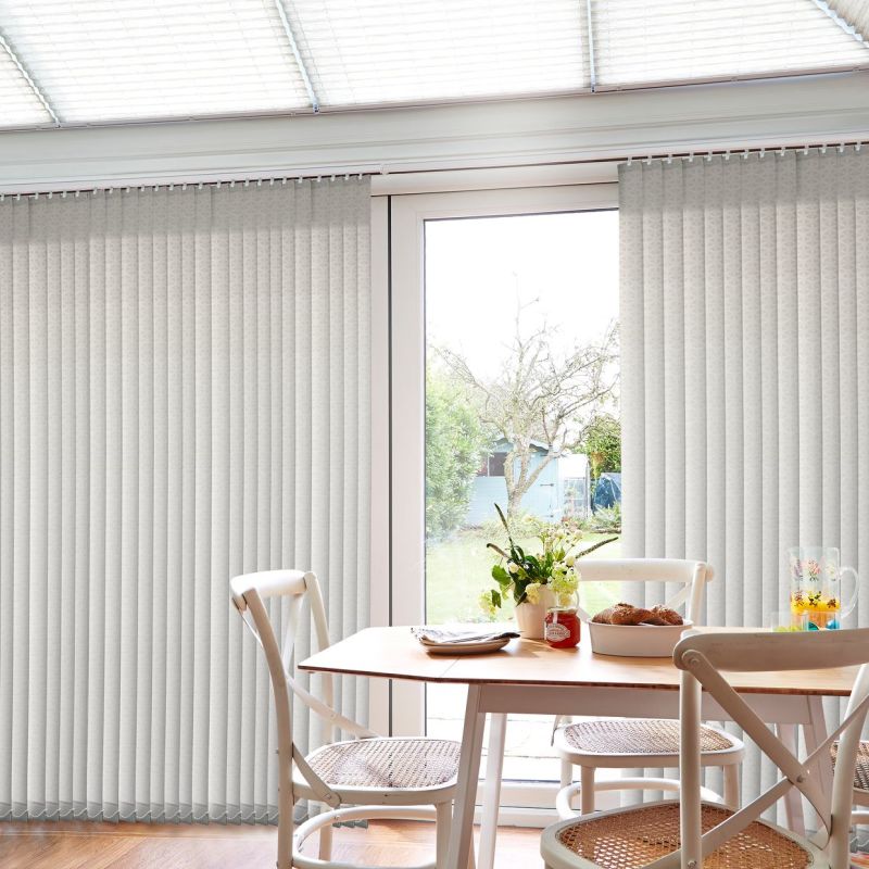 Vertical pale beige blinds, full‑height and mostly closed with a central gap, filtering daylight and partially open to a garden view; dining table, chairs and vase of flowers in room.