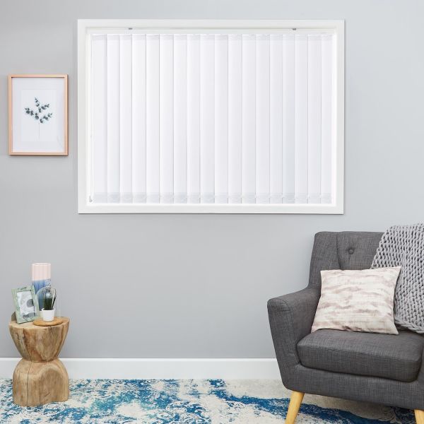 White vertical blinds covering a window, fully closed with scalloped bottom tabs, filtering daylight; in a gray-walled living room with gray armchair, knitted throw, wooden side table and blue rug.