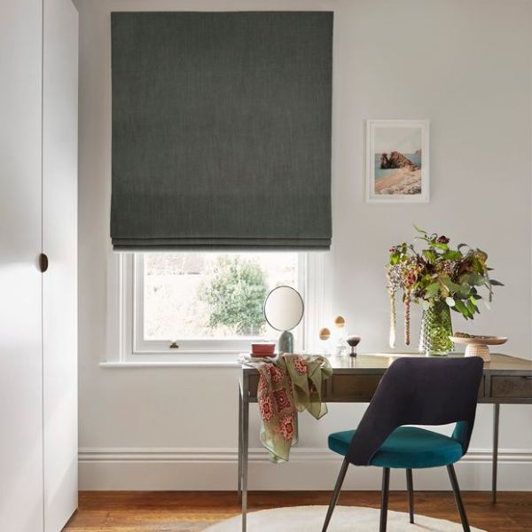 Object: dark grey Roman blind lowered over a window, fabric with a small gap at bottom. Action: partially closed, filtering daylight. Context: home desk vignette with chair, mirror, vase of flowers.