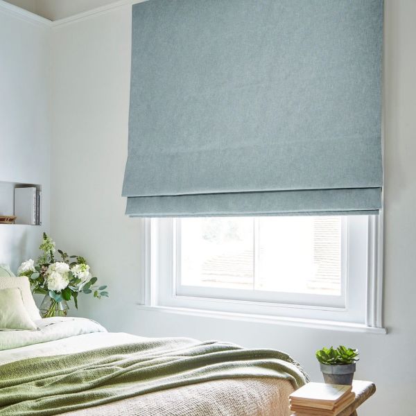 A pale blue Roman shade, lowered with horizontal folds, covers the window, filtering daylight; bedroom with white walls, green bedding, vase of white flowers and a small succulent.