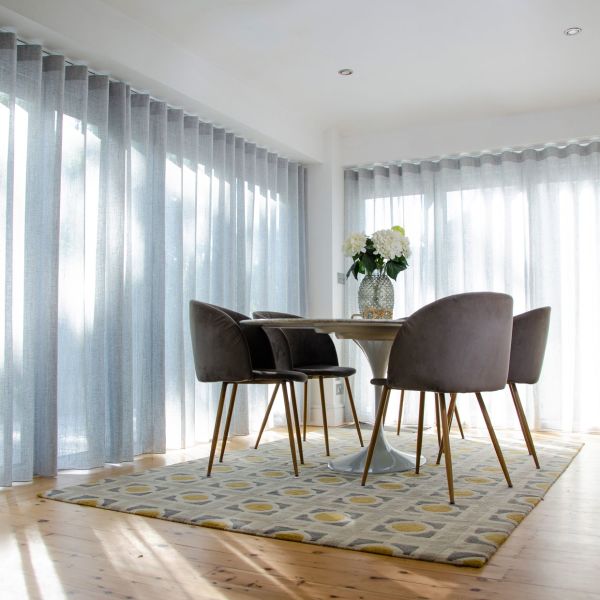 Object: sheer floor-to-ceiling pleated curtains on two walls, pale gray with vertical folds. Action: drawn closed, softly filtering daylight. Context: sunlit dining nook with round table, four chairs, patterned rug.