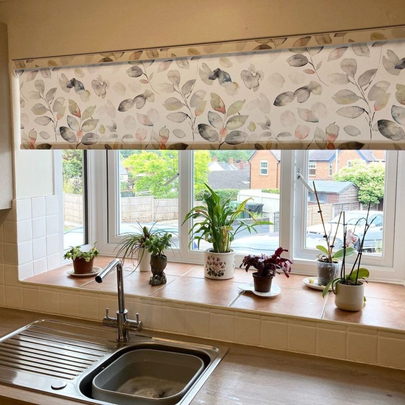 Floral roller blind, lowered across a three-panel window and patterned with watercolor leaves, filtering daylight over a kitchen sink and tiled sill with potted plants, suburban houses visible beyond.