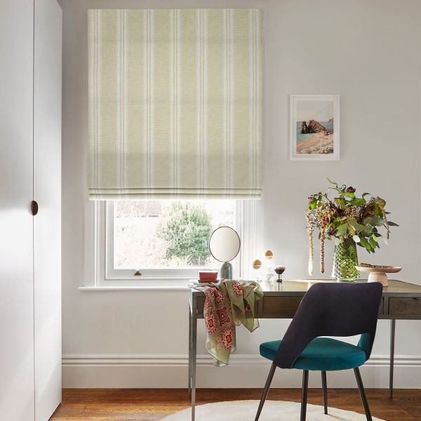 Striped beige Roman blind lowered over window with subtle white vertical stripes, filtering soft daylight, folded at the bottom; home study with desk, teal chair, mirror and vase of flowers.