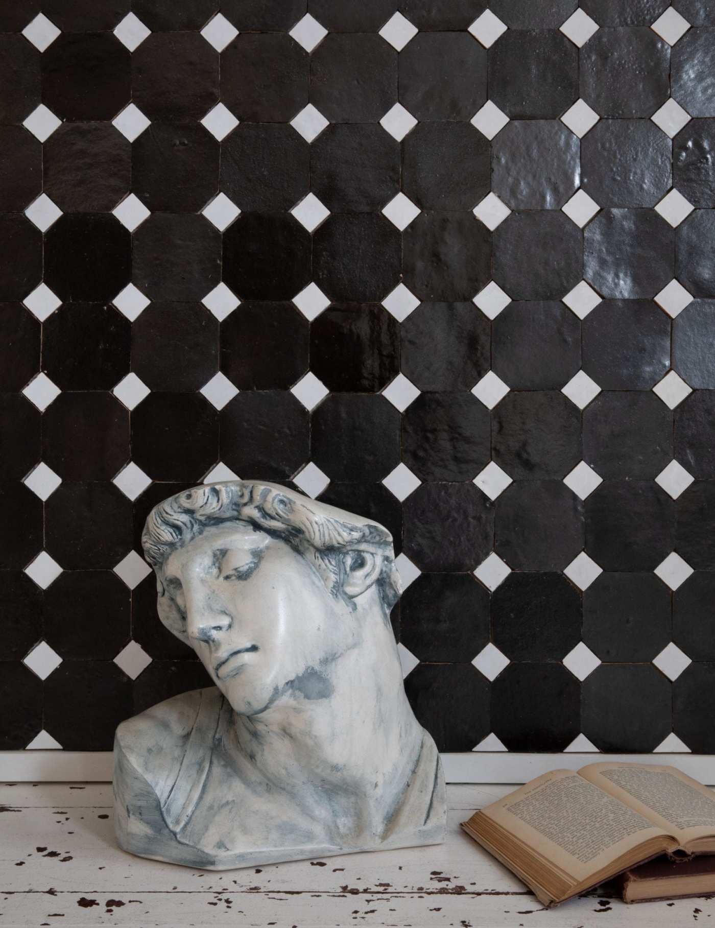 a bust on a table next to a book against a black and white tile wall.