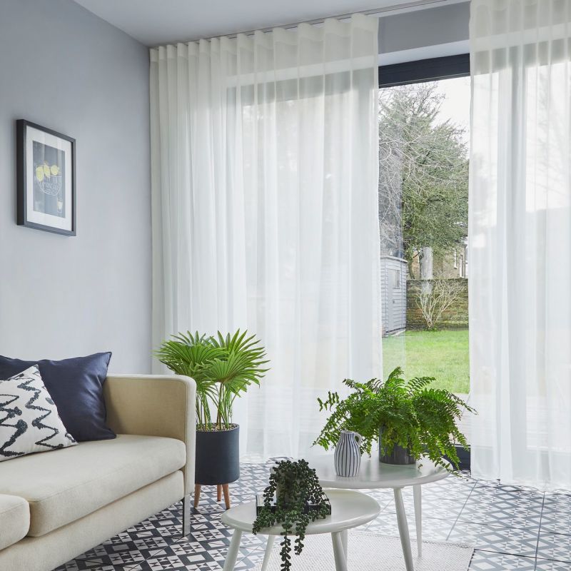 Sheer floor-to-ceiling pleated curtains, partially drawn, softly filter daylight through sliding glass doors into a living room with a pale sofa, patterned tile floor, potted plants, and a lawn view.