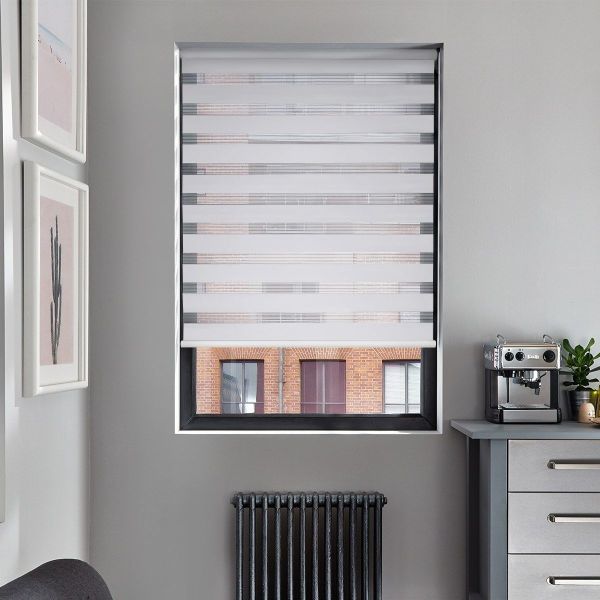 Striped dual-layer roller blind, alternating opaque and sheer bands, partially lowered filtering light, revealing lower window view of a brick building; set in a grey kitchenette with espresso machine, radiator.