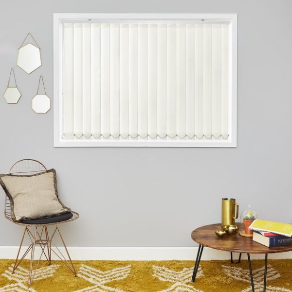 Cream vertical blinds (inside window frame, scalloped weighted bottoms) — closed, softly diffusing daylight — in a grey-walled living room with a wire chair, round wooden coffee table, and mustard patterned rug.