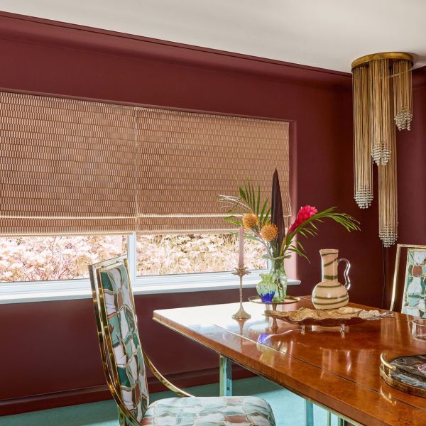 Beige woven Roman shades, fully lowered with visible horizontal folds, filtering soft daylight through the window; burgundy dining room with a wooden table, floral vase, patterned chairs, brass chandelier.