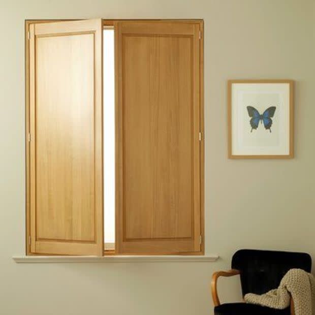 Interior oak shuttersâ€”two hinged panelsâ€”cover a rectangular window; one panel is slightly ajar, admitting bright light into a neutral-toned room with a framed blue butterfly print and a black armchair.