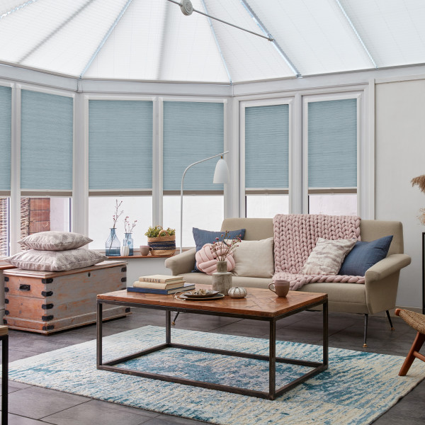 Light-blue pleated cellular blinds, lowered over tall conservatory windows with uncovered bottoms; filtering soft daylight; in a bright, cozy living area with beige sofa, wooden coffee table and rug.