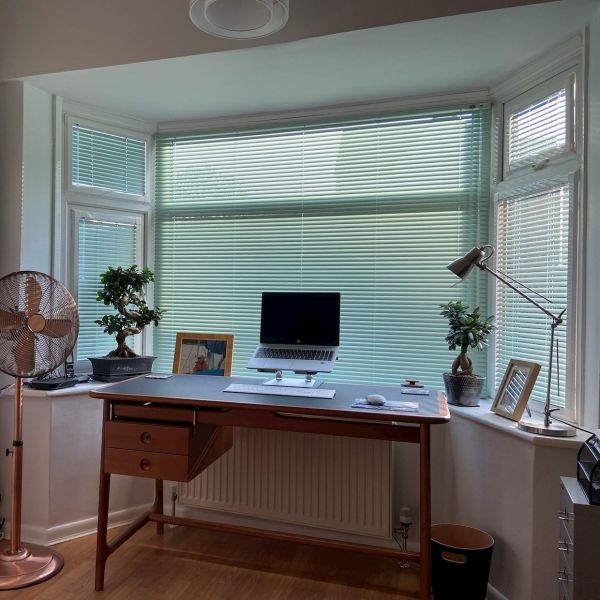 Horizontal pale green Venetian blinds covering a three-panel bay window, mostly closed, filtering soft daylight into a tidy home office with wooden desk, laptop, adjustable lamp, and potted plants.