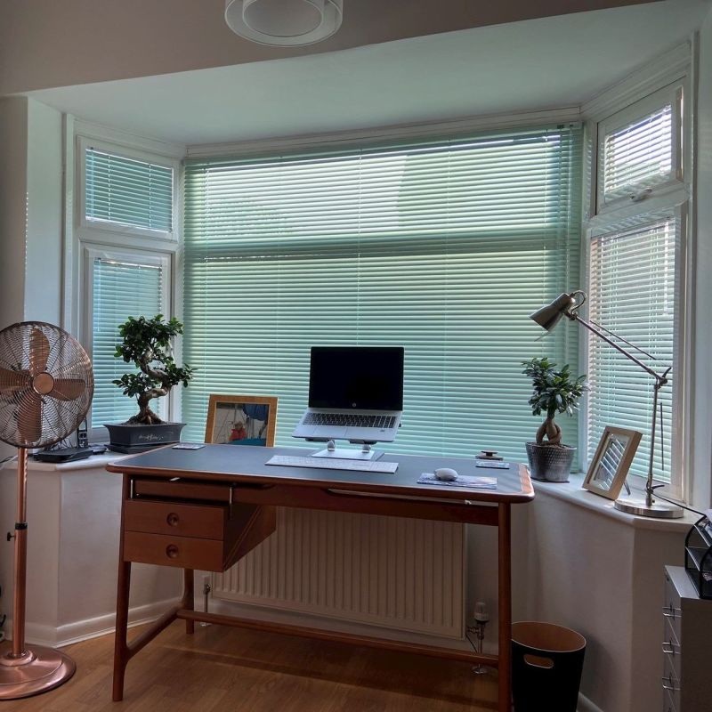 Horizontal pale green Venetian blinds covering a three-panel bay window, mostly closed, filtering soft daylight into a tidy home office with wooden desk, laptop, adjustable lamp, and potted plants.