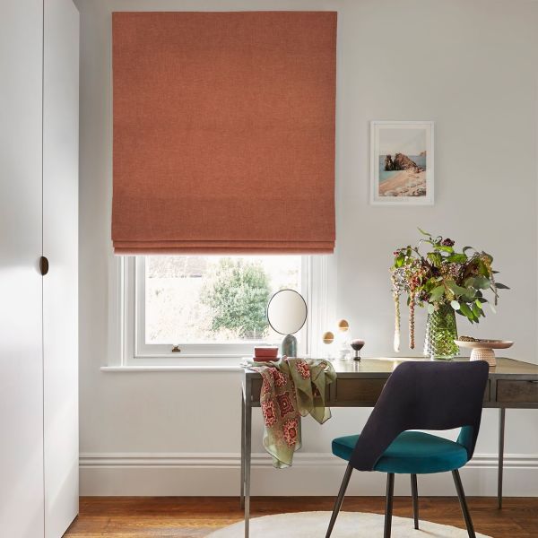 Terracotta fabric Roman blind, fully lowered with horizontal folds at bottom, filtering soft daylight through the window; small desk with mirror, teal chair and vase of flowers in a bright neutral study.