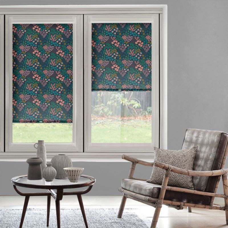 Floral-patterned roller blinds cover two tall windows—left fully lowered, right half-lowered—featuring teal and pink botanicals; they filter daylight into a gray living room with wicker chair and ceramic tableware.