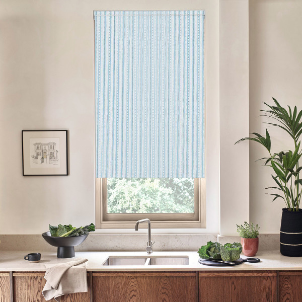 Blue-striped roller blind covers a kitchen window above a stainless-steel faucet; light filters through. On the countertop: a bowl of greens, potted herbs, folded towel, framed sketch and tall plant.
