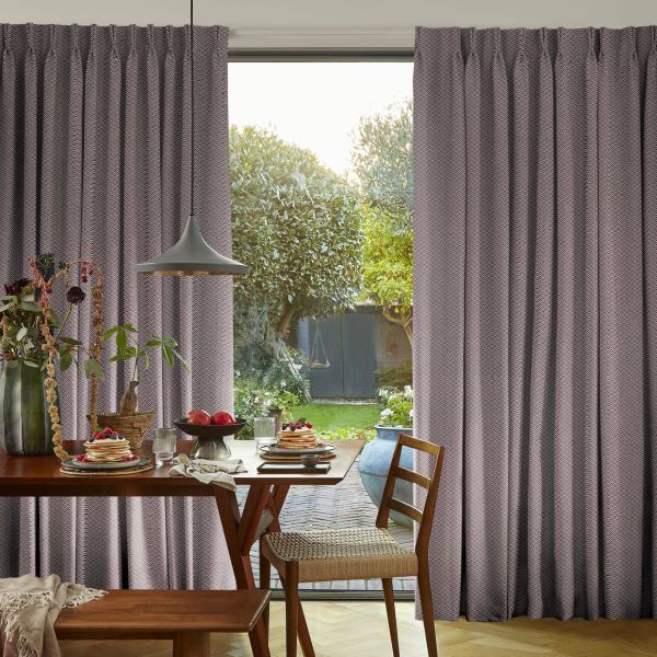 Floor-length pleated patterned curtains, pulled to sides with a central opening, framing a glass door; filtering daylight into a dining area with wooden table set for breakfast and garden beyond.