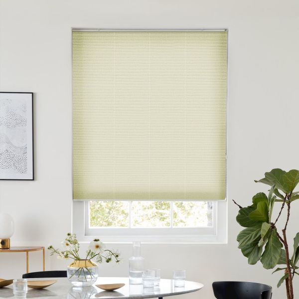 Light-yellow pleated cellular shade, fully lowered over a sash window, filtering daylight; set in a bright minimalist dining area with round table and vase, framed art left, fiddle‑leaf fig right.