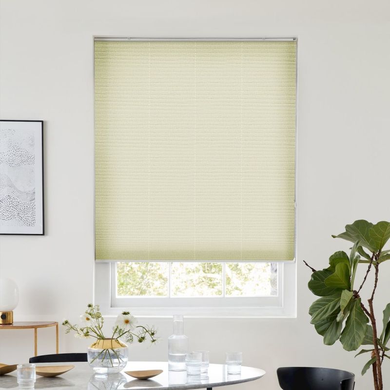 Light-yellow pleated cellular shade, fully lowered over a sash window, filtering daylight; set in a bright minimalist dining area with round table and vase, framed art left, fiddle‑leaf fig right.