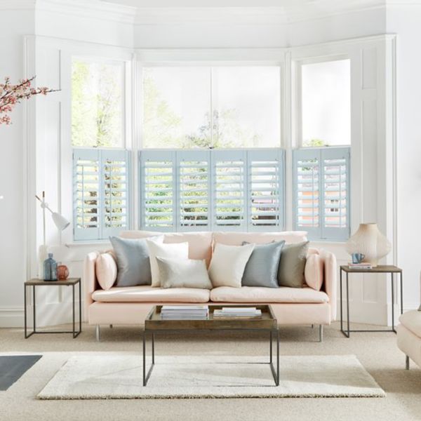 Pale-blue café-style plantation shutters cover the lower halves of a bay window; slatted panels partially open, filtering daylight into a bright living room with pastel sofa, cushions and coffee table.
