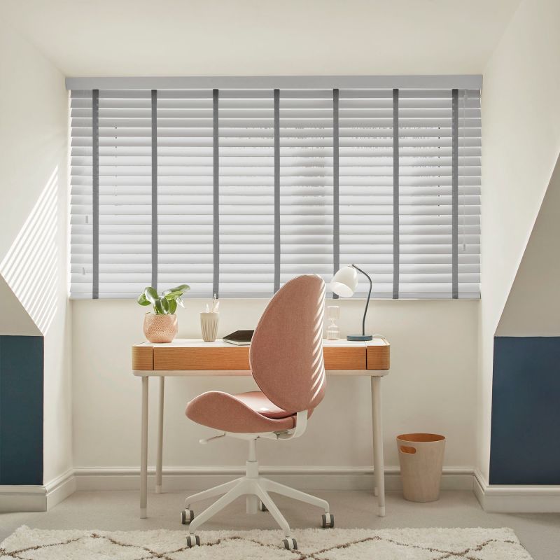 White horizontal slatted blinds with grey vertical tapes cover a wide dormer window, closed and filtering soft daylight; small home study with wooden desk, pink swivel chair and lamp.