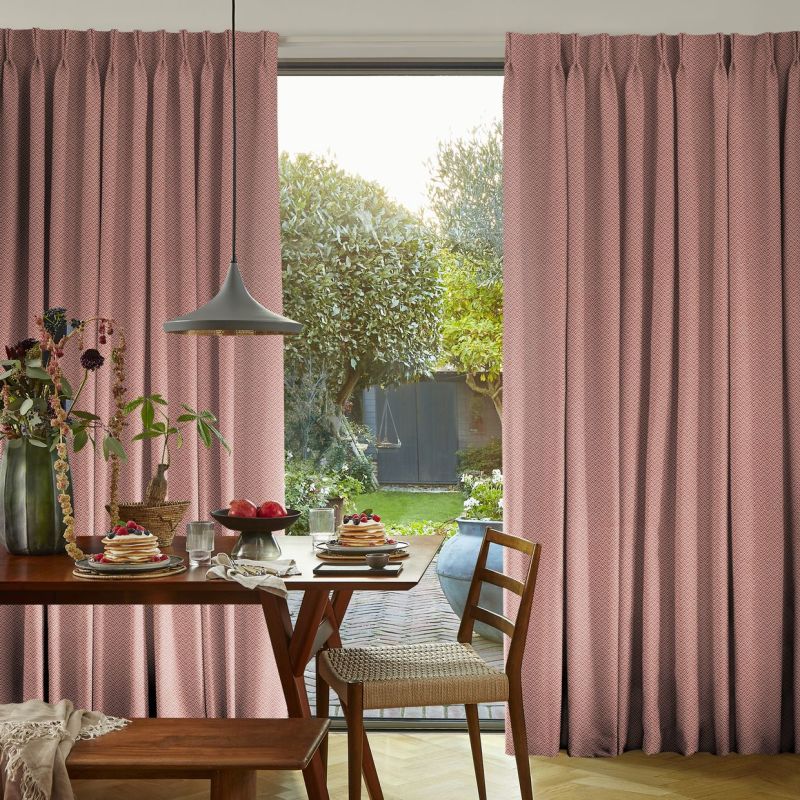 Pink chevron-patterned full-length curtains, parted centrally to reveal a sunlit garden; filtering soft daylight into a dining nook with wooden table, pendant lamp, breakfast plates, and greenery.