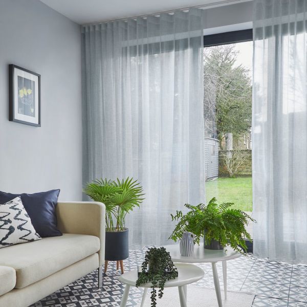 Sheer pale-gray floor-length curtains hung from ceiling track, softly pleated and partially drawn, filtering daylight; living room with beige sofa, patterned tiled floor, nesting white tables, potted plants, garden view.