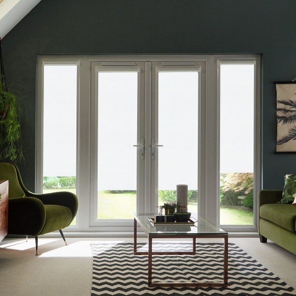 French glass doors remain closed, admitting bright daylight into a living room furnished with green upholstered chairs, a glass coffee table on a black-and-white chevron rug, and potted plants.