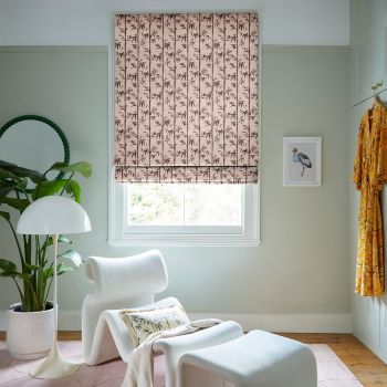 Pink Roman blind patterned with dark bamboo, lowered and softly filtering daylight, covers a window in a pale-green sitting room with a white lounge chair, lamp, plant, hanging robe.