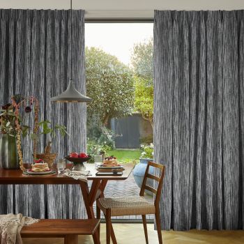 Full-length grey textured curtains with vertical striations, parted at center; framing and filtering sunlight onto a wooden dining table with pancakes and fruit, lamp and chairs, opening onto a garden.