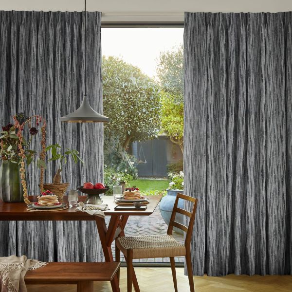 Full-length grey textured curtains with vertical striations, parted at center; framing and filtering sunlight onto a wooden dining table with pancakes and fruit, lamp and chairs, opening onto a garden.
